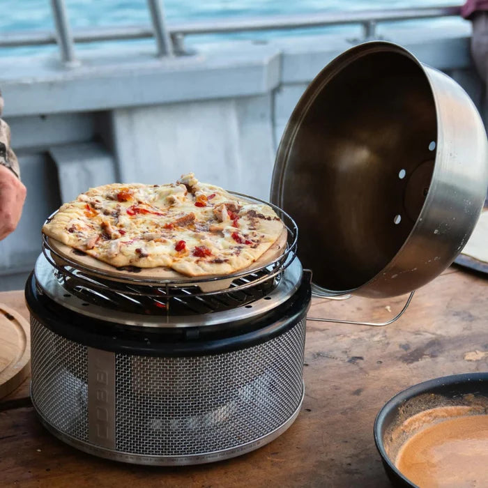 Portable pizza oven with a pizza inside on a wooden surface by the sea.
