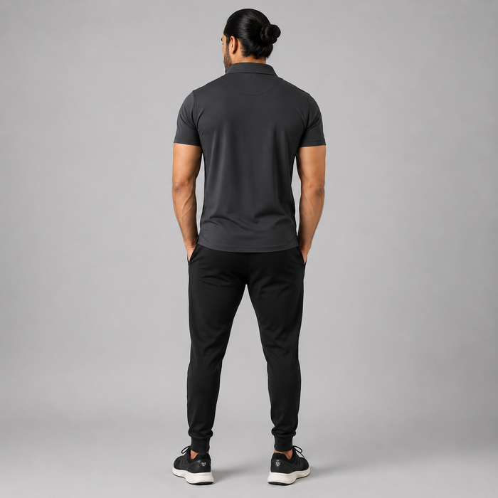 A man viewed from behind wearing a dark gray short-sleeve polo shirt, black jogger pants, and black sneakers, standing against a solid gray studio background.