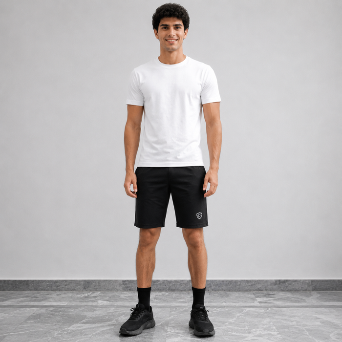 A man modeling a white crew-neck t-shirt and black athletic shorts with a small white shield logo on the left leg, standing against a light gray studio wall.