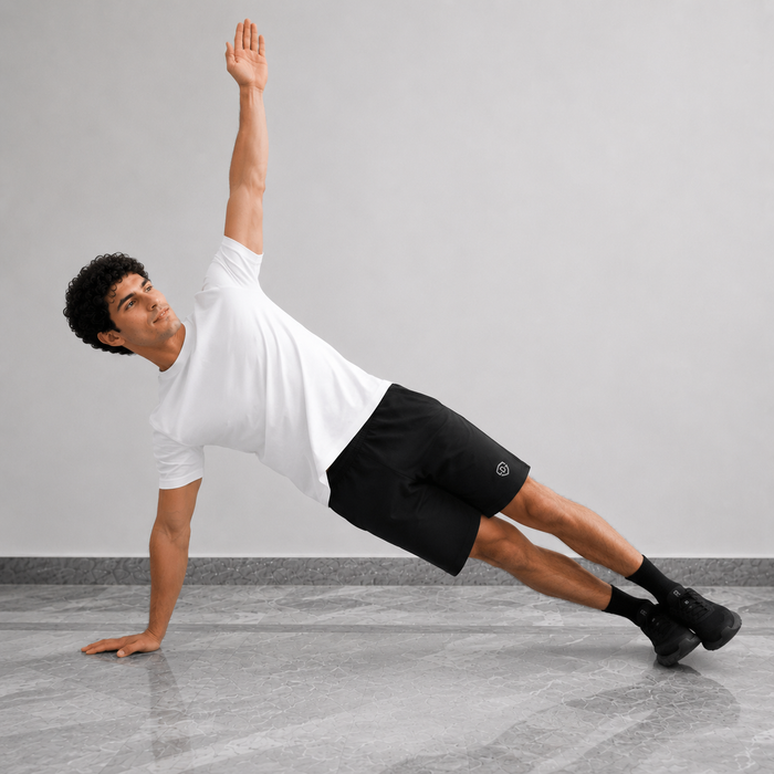 A man in a white t-shirt and black athletic shorts performs a side plank exercise on a gray floor against a light gray wall.