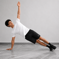 A man in a white t-shirt and black athletic shorts performs a side plank exercise on a gray floor against a light gray wall.