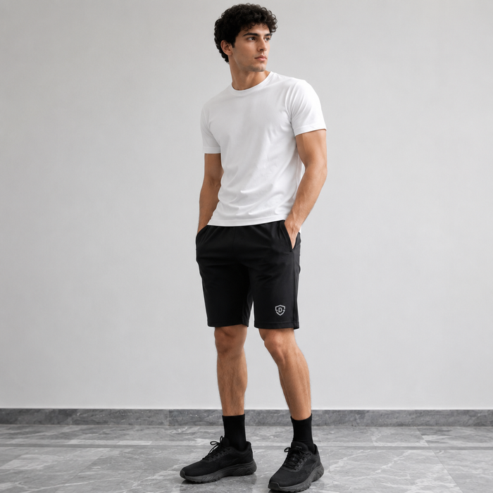 A man modeling a white crew-neck t-shirt and black athletic shorts with a small shield logo on the left leg, paired with black socks and black sneakers, standing against a light gray studio wall.