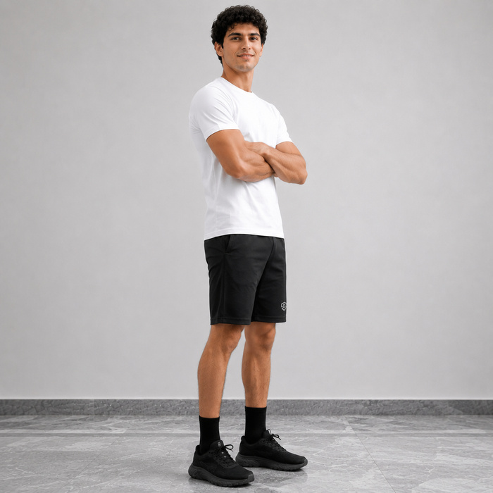 A man stands in a studio wearing a white short-sleeve t-shirt, black athletic shorts, black socks, and black sneakers against a light gray background.