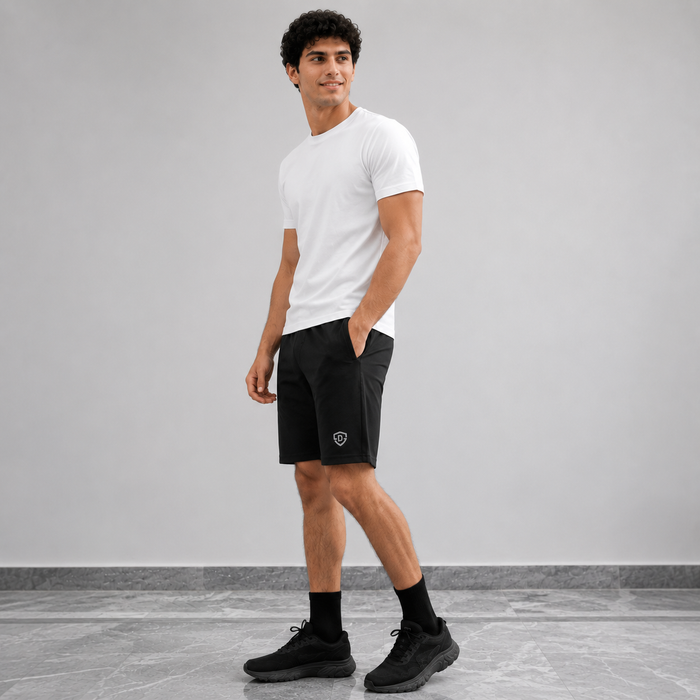 A man wearing a white crew-neck t-shirt, black athletic shorts with a small shield logo on the left leg, black socks, and black sneakers, standing against a light gray wall and tiled floor.