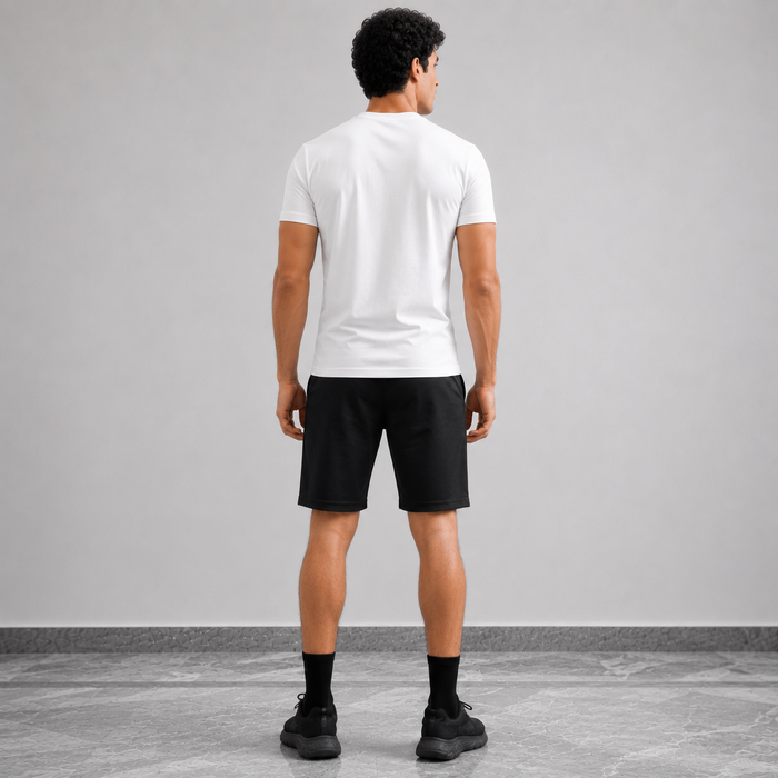 A man viewed from behind wearing a white short-sleeve t-shirt, black athletic shorts, black socks, and black sneakers, standing against a light gray studio wall.