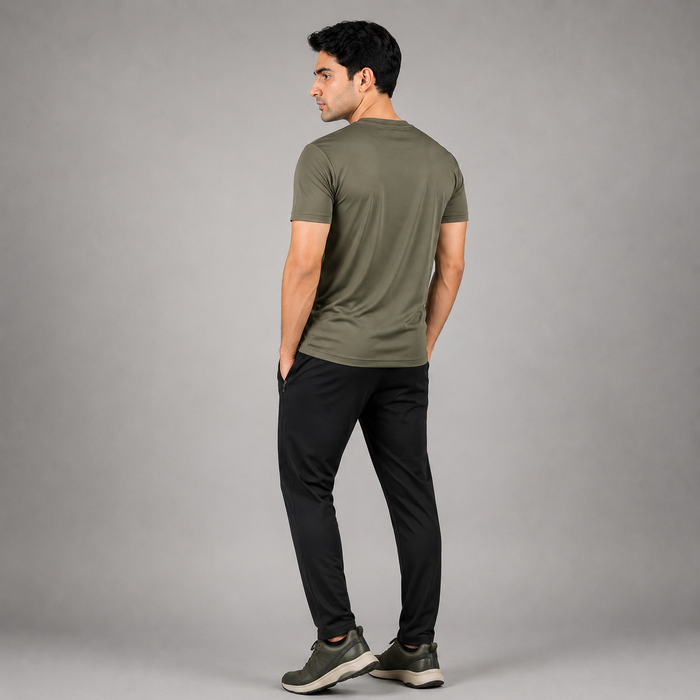 A man wearing an olive green short-sleeve t-shirt and black pants, shown from a rear three-quarter view against a solid gray studio background.