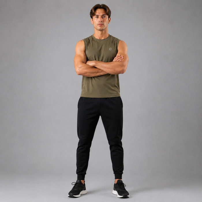 A man stands with arms crossed wearing an olive green sleeveless athletic tank top and black jogger pants against a solid gray studio background.