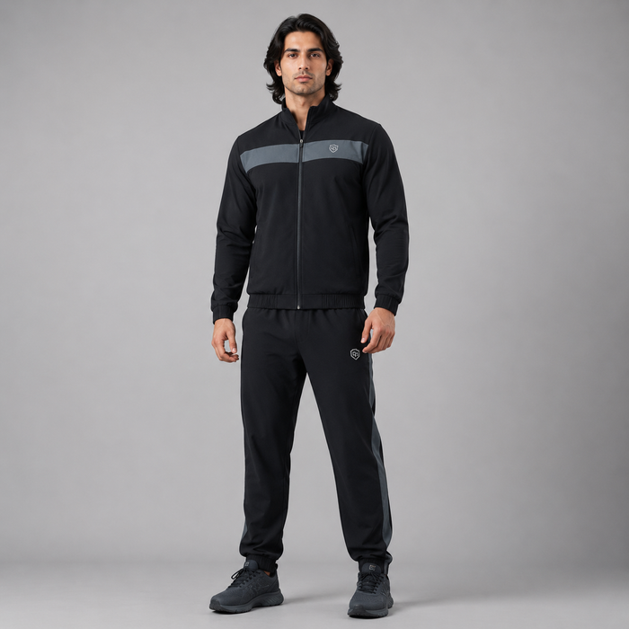 A man modeling a black athletic tracksuit with a gray chest stripe and matching gray side stripes on the pants, photographed against a neutral gray studio background.