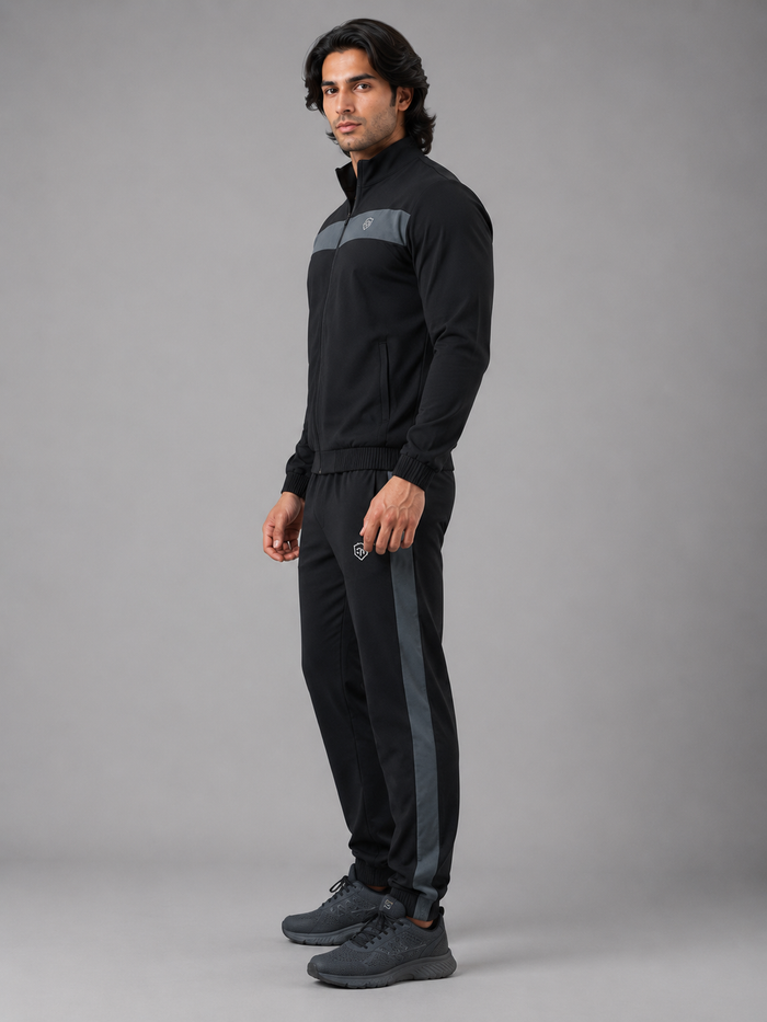 A man modeling a black athletic tracksuit with gray stripe accents on the chest and down the pants, paired with dark gray sneakers, against a solid gray studio background.