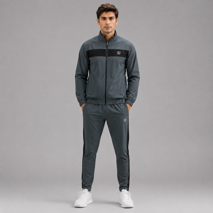 A man modeling a dark gray athletic tracksuit with a black chest stripe and white sneakers, standing against a solid gray studio background.