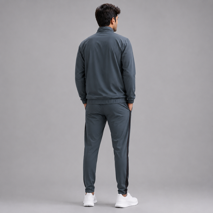 A man viewed from behind wearing a dark gray athletic tracksuit with a zip-up jacket and matching jogger pants, paired with white sneakers, against a solid gray studio background.