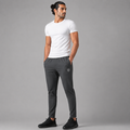 A man modeling dark gray athletic jogger pants with a small shield logo on the left thigh, paired with a white t-shirt and black sneakers, against a solid gray studio background.