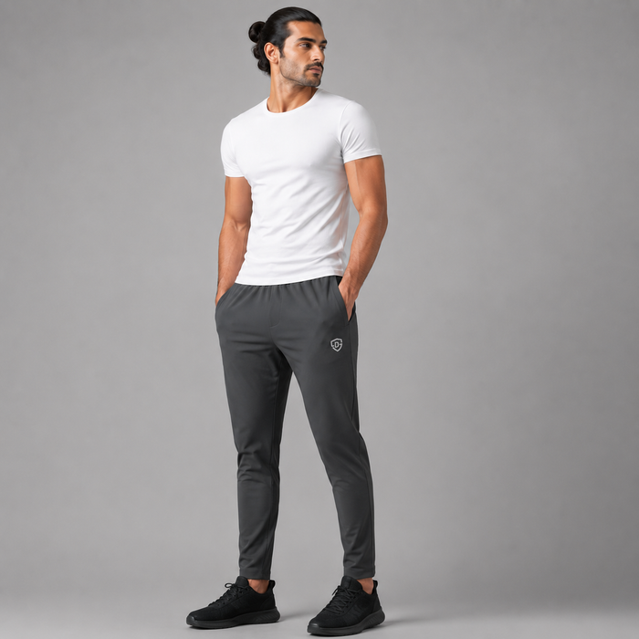 A man modeling dark gray athletic jogger pants with a small shield logo on the left thigh, paired with a white t-shirt and black sneakers, against a solid gray studio background.