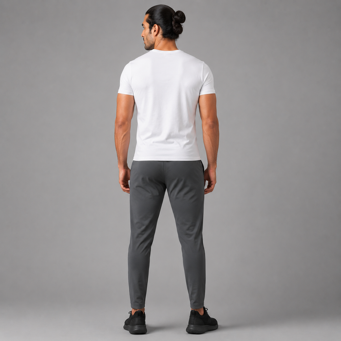 A man viewed from behind wearing Charcoal Grey Trousers, a white short-sleeve t-shirt, dark gray slim-fit pants, and black sneakers, standing against a solid gray studio background.