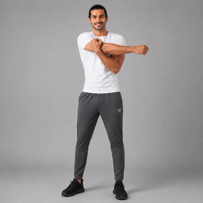 A man modeling dark gray athletic pants and a white t-shirt, stretching his arm while standing against a solid gray studio background.