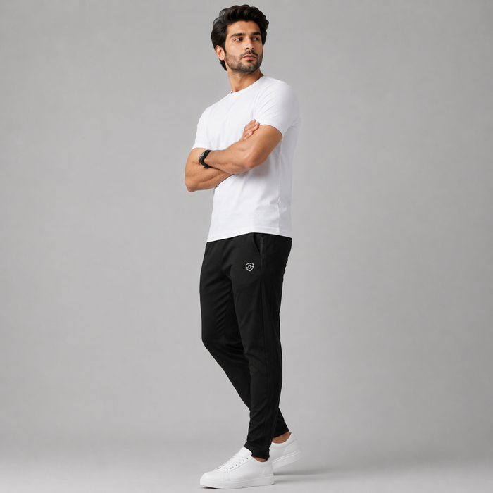 A man modeling a white crew-neck t-shirt and black athletic pants with a small white shield logo on the left thigh, paired with white sneakers, against a solid gray studio background.