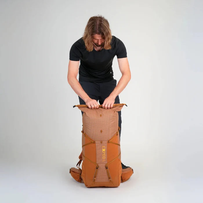 Person opening a brown backpack with 'Exped' branding on a light gray background