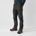 A person standing wearing black Fjallraven trekking trousers with multiple pockets and adjustable straps on the leg endings. They are also wearing a brown boot and a dark green shirt.