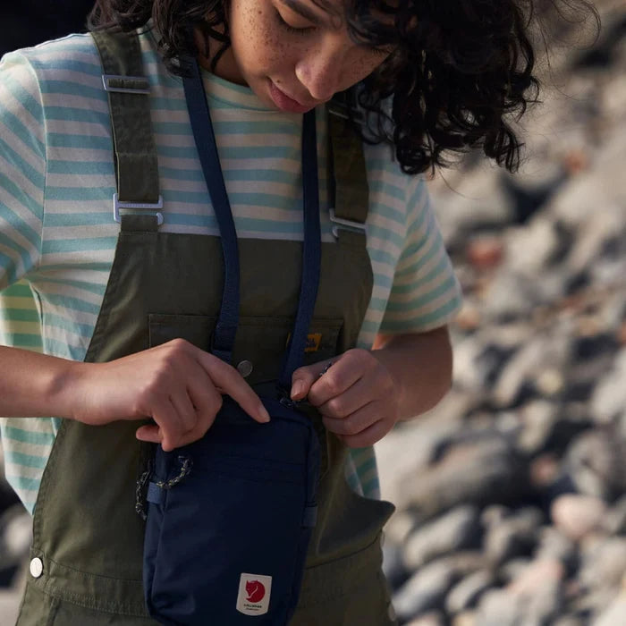 Person wearing a striped shirt and green overalls with a blue bag, standing on a rocky surface.