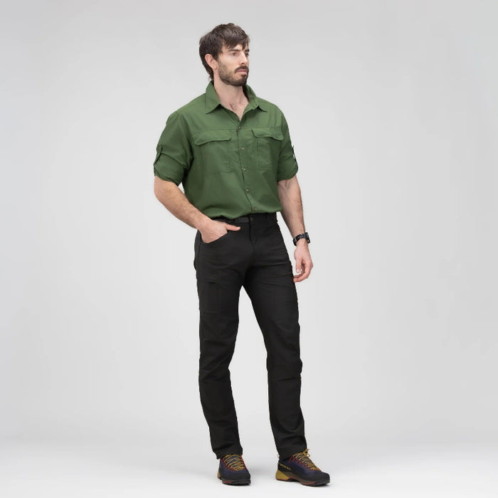 A man wearing an olive green long-sleeve button-up shirt and black pants, standing against a light gray studio background.