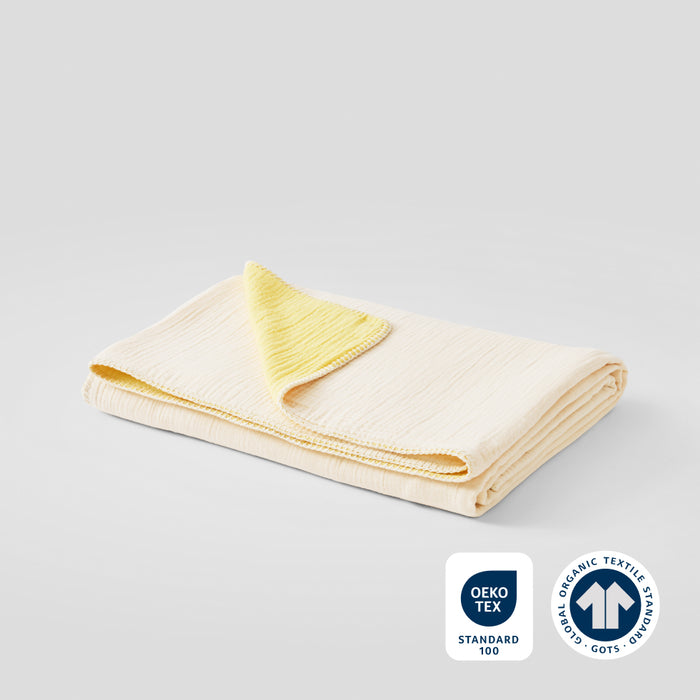 A folded lemon icing cotton muslin blanket with OEKO-TEX and GOTS certification badges in the lower right corner.