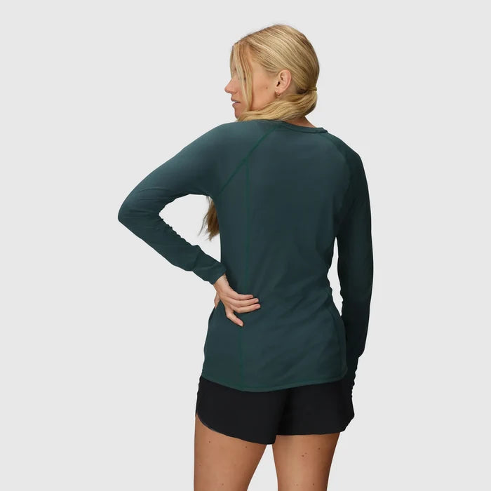 Woman wearing a green long-sleeve outdoor research long sleeve hiking top and black hiking shorts on a light gray background