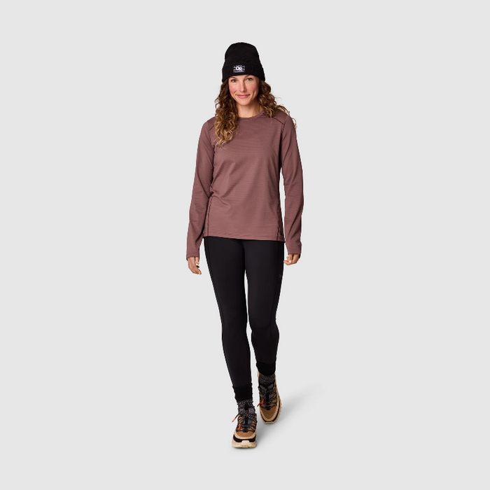 A woman modeling a long-sleeve athletic top in a dusty rose color, paired with black leggings, a black beanie, and hiking boots, against a light gray studio background.