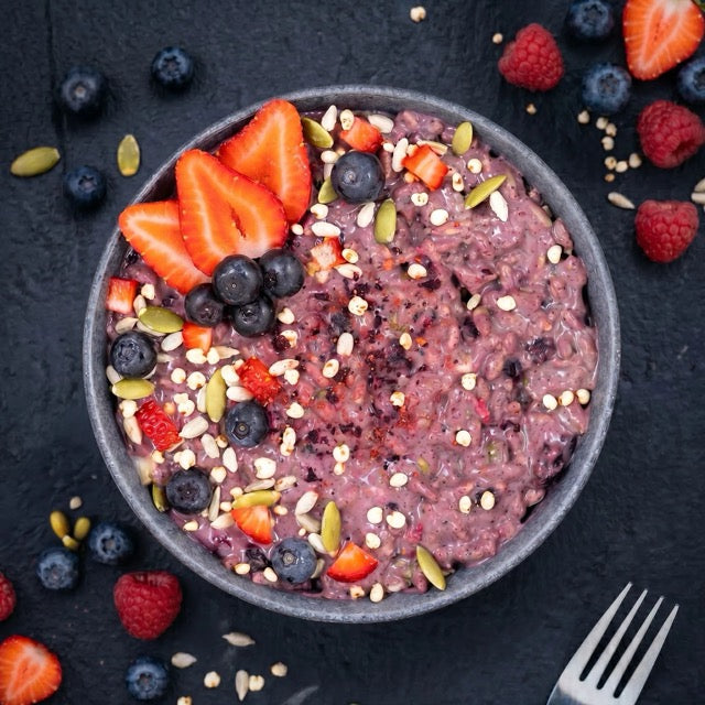 Bowl of purple porridge with berries and seeds on a dark surface
