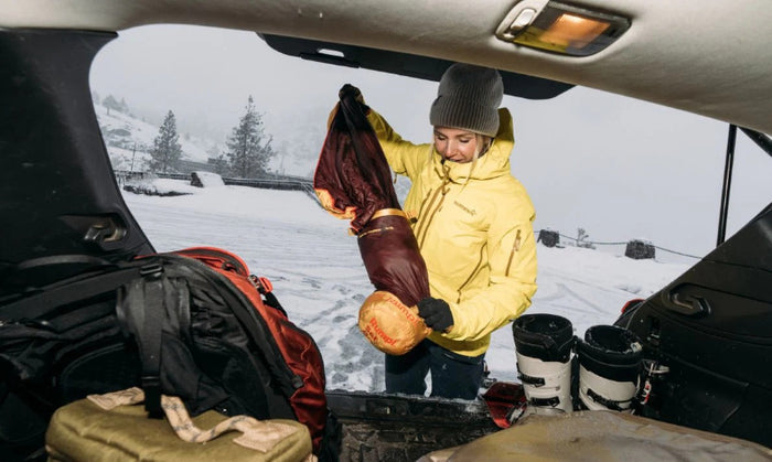 A person in a yellow jacket and gray beanie unpacks a brown camping sleeping bag from the trunk of a car, with a snowy outdoor landscape in the background.
