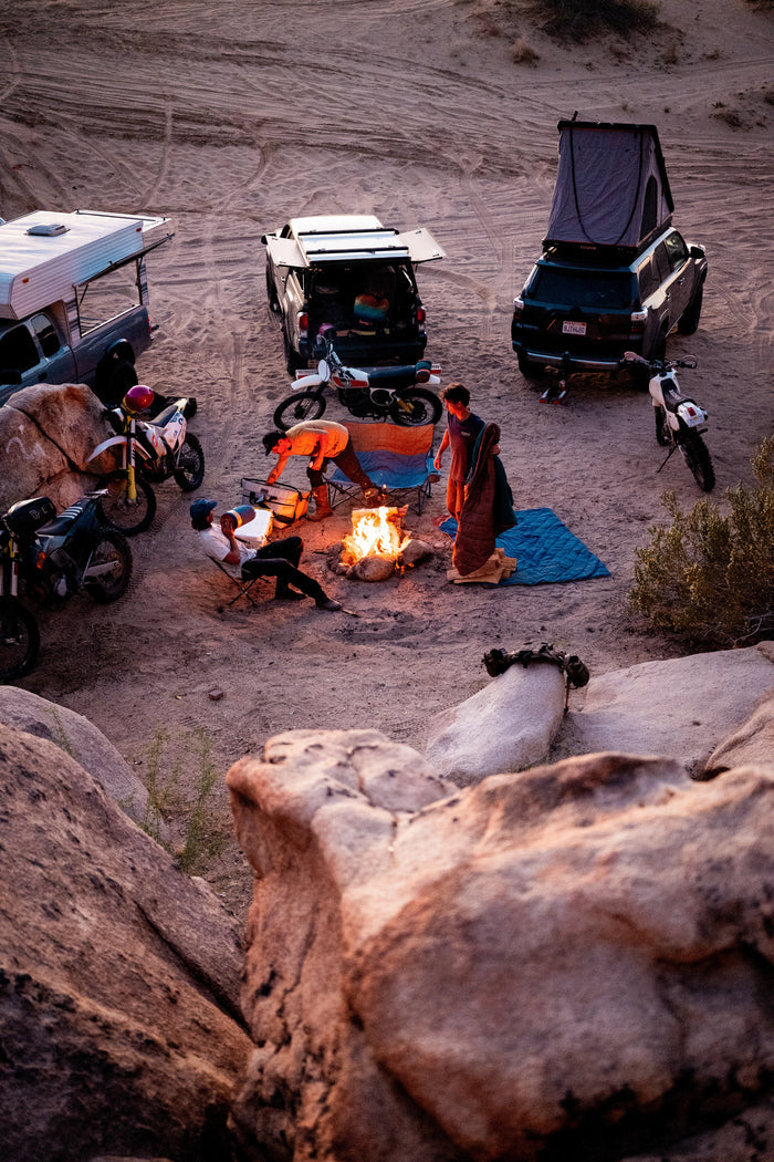 Group of people around a campfire in a desert setting with vehicles and camping gear.