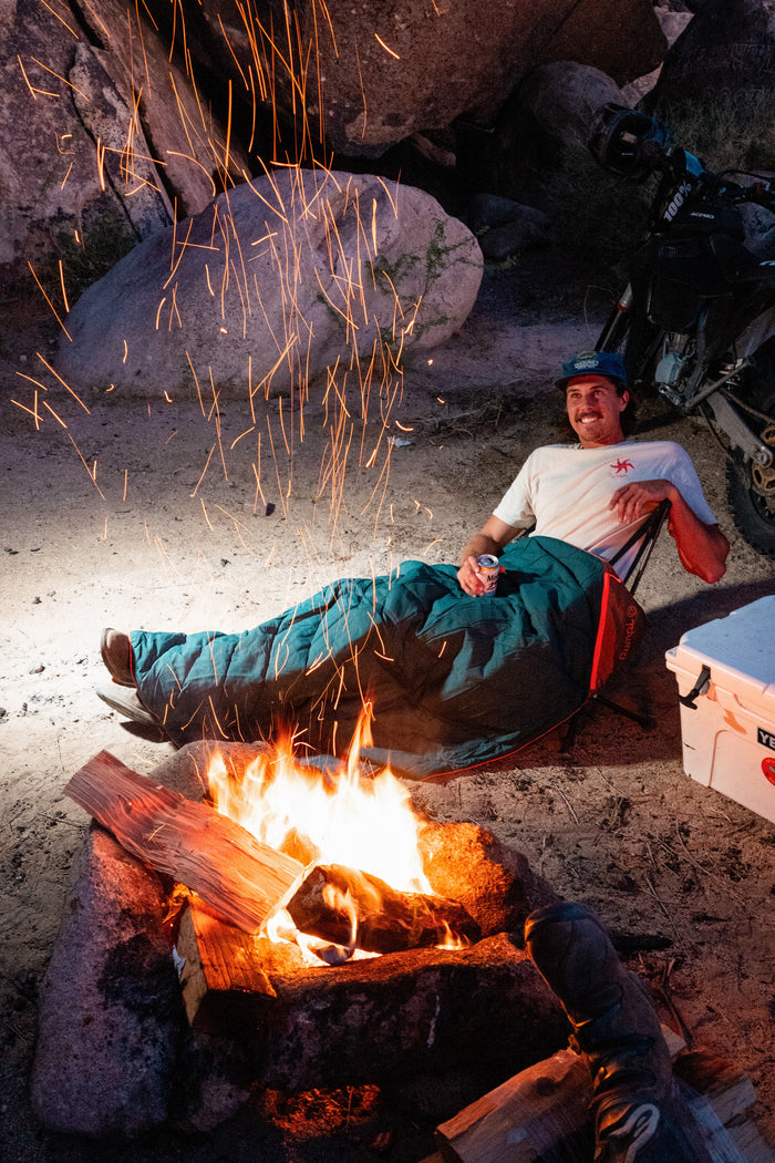 Person sitting by a campfire with sparks flying, near a cooler and rocks.