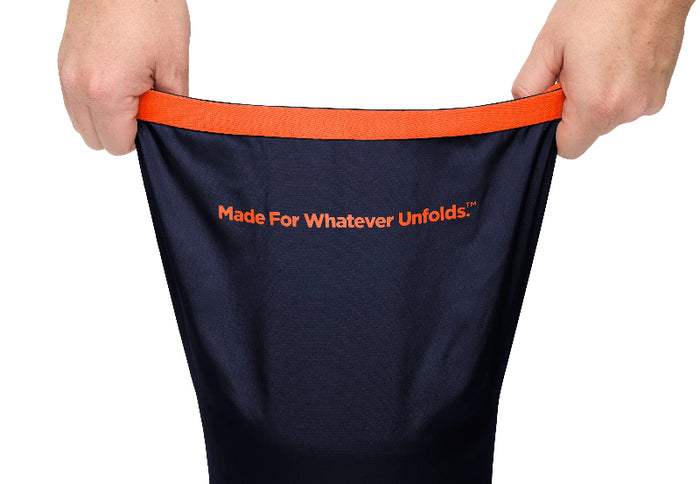 A navy blue bag with an orange waistband and orange text reading 'Made For Whatever Unfolds.' is being held open by two hands against a white background.