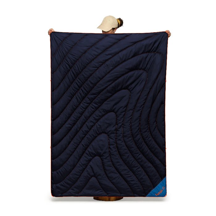 A person wearing a beige cap holds up a large quilted navy blue sleeping pad with wavy contour stitching against a plain white background.