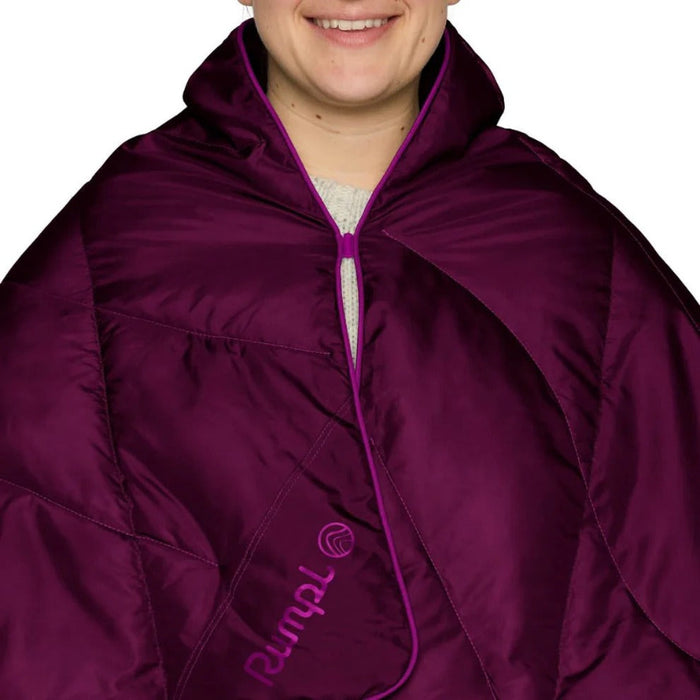 A close-up of a person wearing a deep purple puffer poncho or cape with a matching purple zipper and trim. The brand name 'Rumpl' is printed in purple on the lower left side.