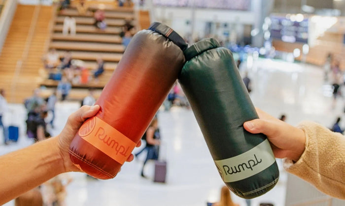 Two people holding compressed inflatable sleeping bags in a public indoor space. One bag is reddish-brown with an orange label reading Rumpl, and the other is dark green with a white label reading Rumpl.