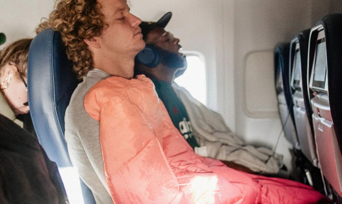 A person sitting in an airplane seat wrapped in a bright coral-pink sleeping bag or blanket, with another passenger reclining in the seat beside them.