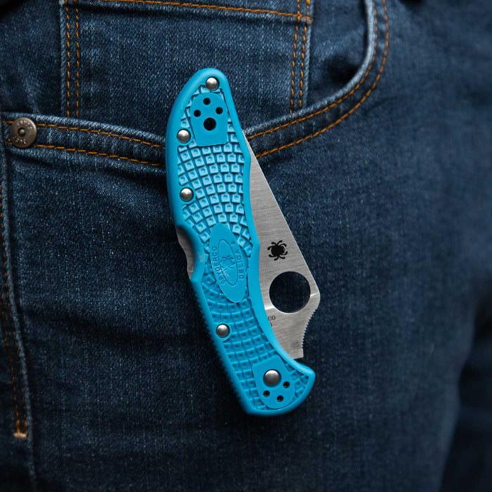 Blue folding knife with textured handle and metallic blade peeking out from a blue denim pocket.